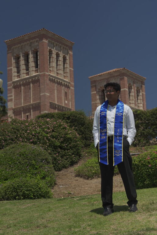 UCLA graduate standing on campus between the towers in a blue stole.